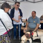 Rendezvénysátorban két egyesület tag beszélget, egyik a padon ül és előtte ott van Vackor a vakvezető kutya. Másik kezében egy fehér bot.